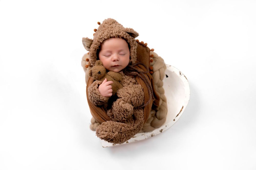 Newborn wearing brown bear outfit sleeping in white egg-shaped prop.