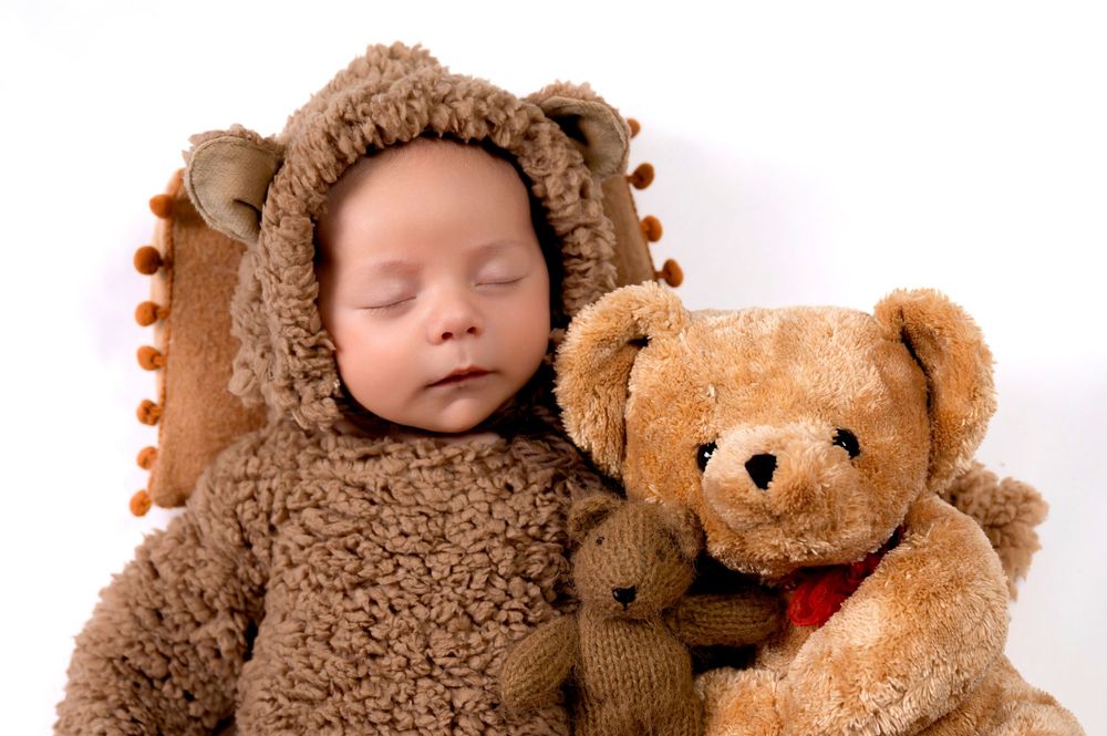 Sleeping baby dressed in plush brown bear costume next to matching teddy bear.