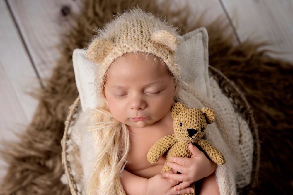 Sleeping newborn wearing knit bear hat holding tiny teddy bear on soft blanket.