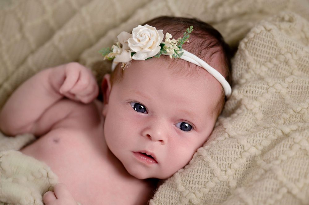 Infant lying on textured cream fabric wearing a delicate floral headband with white roses.