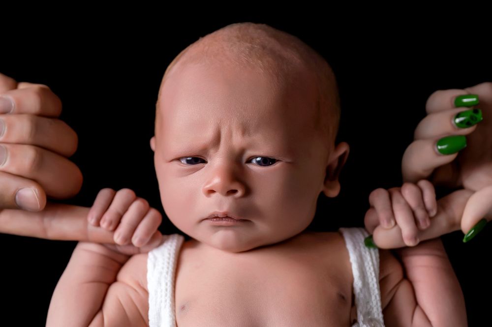 Grumpy newborn baby portrait held up by hands with green nails against dark background.