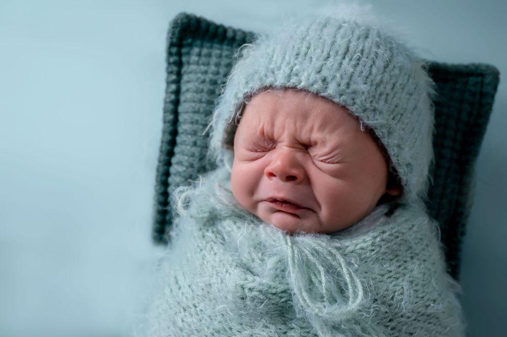 Crying newborn wrapped in gray knit blanket and hat against blue background.