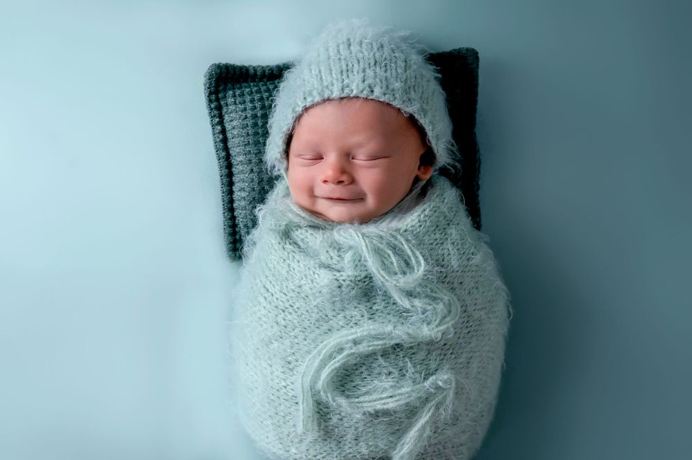 Newborn wrapped in soft blue knit swaddle smiling while lying on dark backdrop.