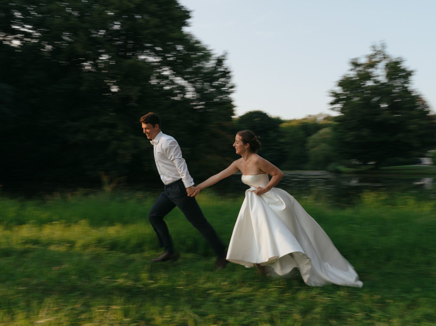 A wedding couple runs playfully through a green field at dusk with their wedding attire in motion.