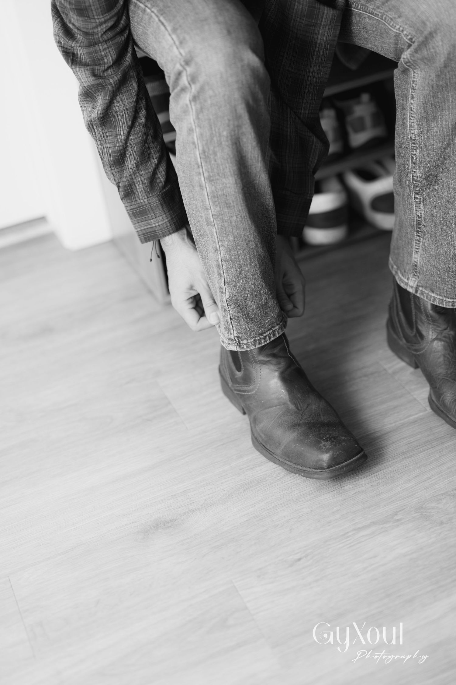Black and white photo of person sitting wearing jeans and boots on wooden floor.