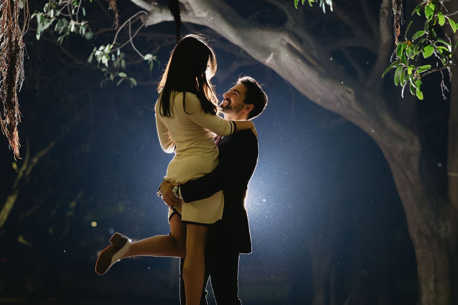 A couple embraces and kisses in a romantic nighttime garden setting with dramatic backlit lighting.
