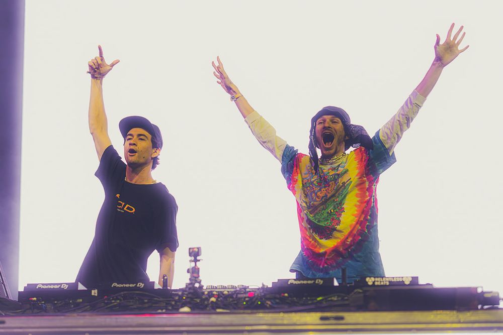 Two DJs performing on stage with raised arms, one wearing a tie-dye shirt and one in dark clothing, at an EDM concert.
