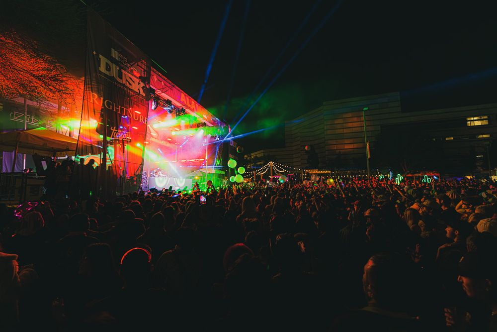 Colorful concert lights illuminate a dark venue with vibrant red and green hues over a large crowd.