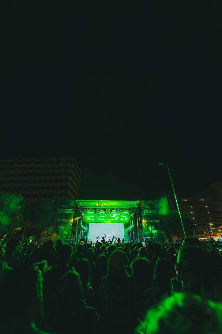 Large concert crowd silhouetted against a green-lit outdoor festival stage at night.