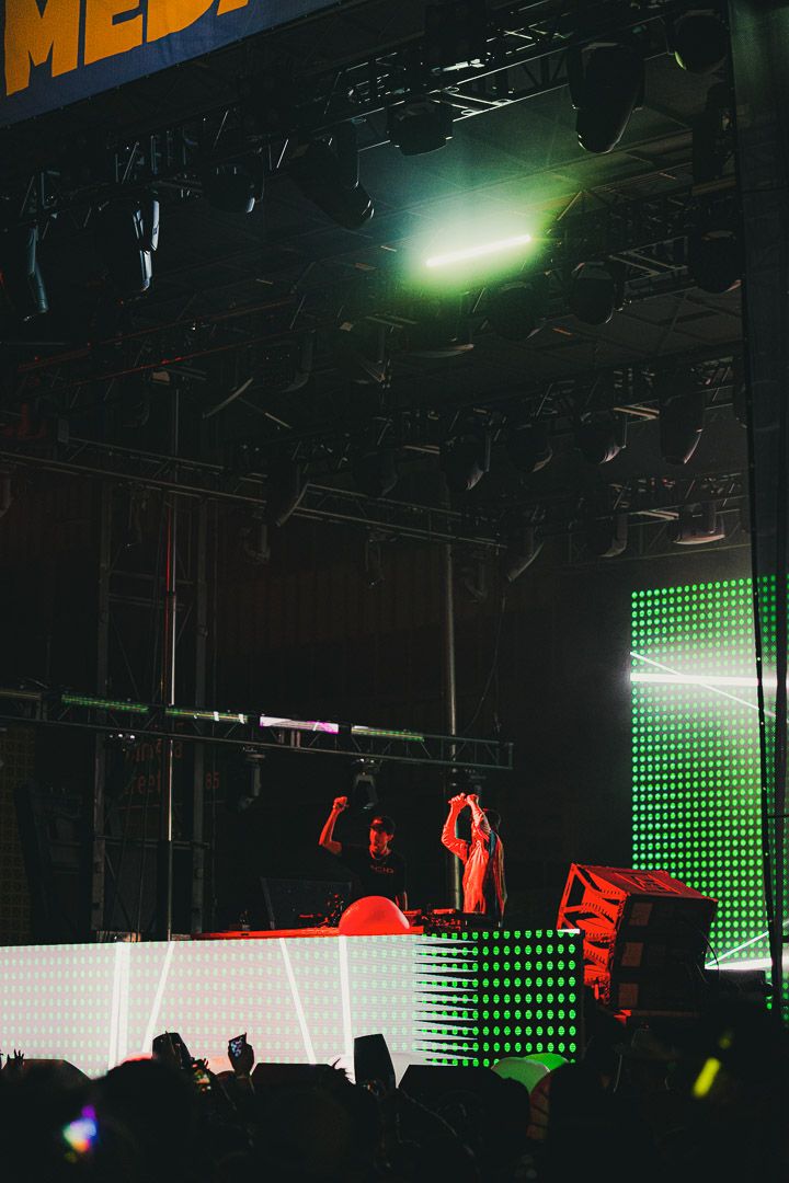 Concert stage setup with dark silhouettes against a green LED wall display and red lighting effects.