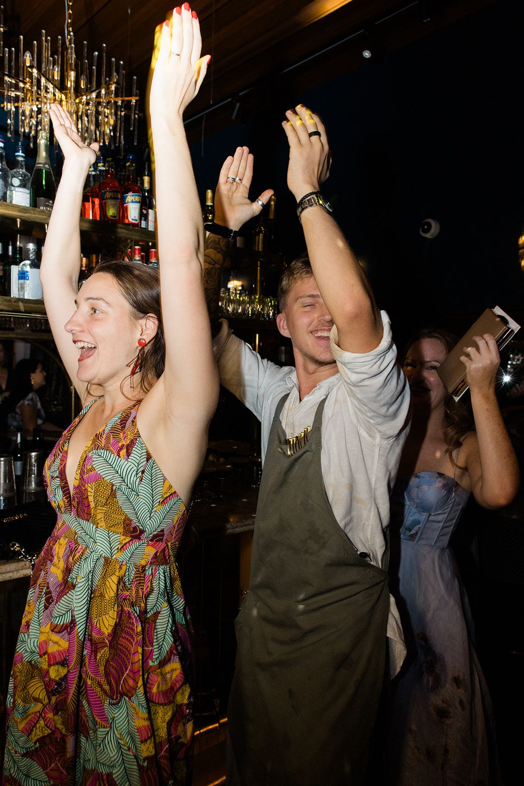 People dancing and raising their hands with excitement in a dimly lit bar or nightclub setting.