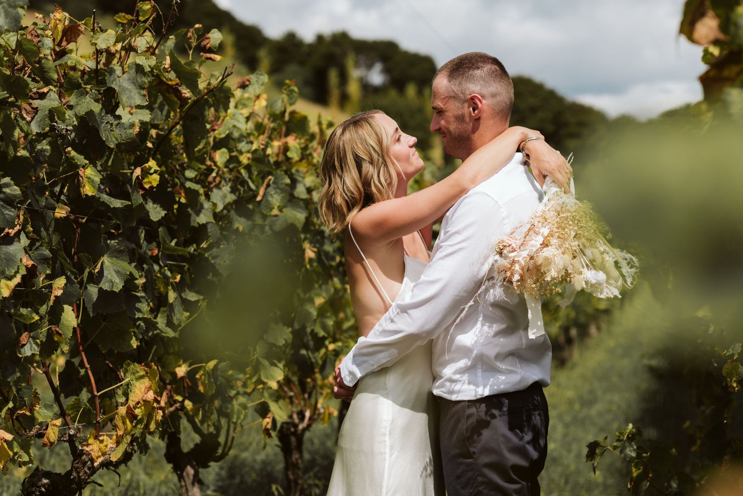 A couple in white and pink attire poses joyfully in a green outdoor setting on a cloudy day.