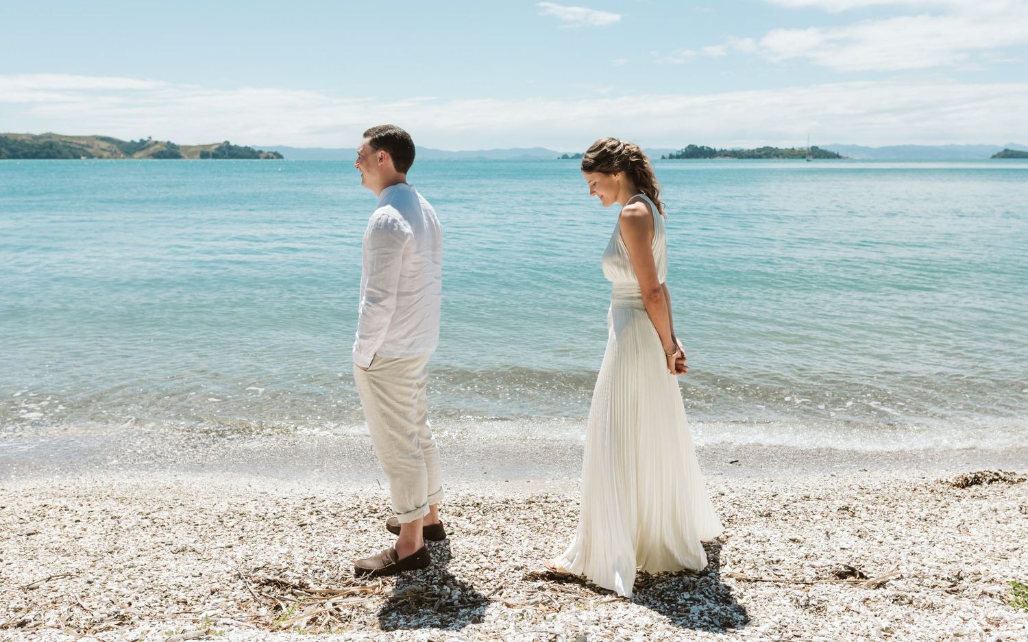 A couple in white and pink attire poses joyfully in a green outdoor setting on a cloudy day.