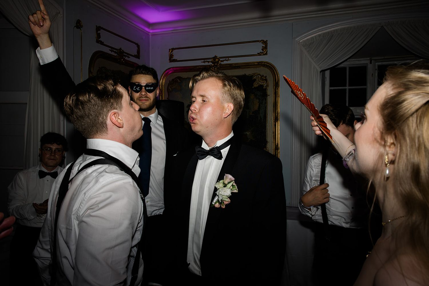 Wedding guests dancing and celebrating on a dance floor under purple lighting at a reception party.
