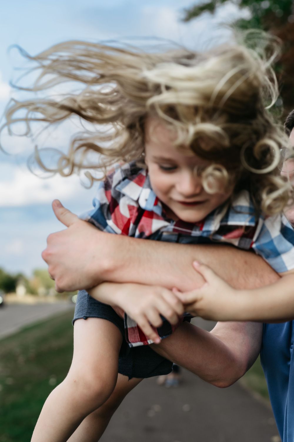 Child with flowing blonde hair being playfully swung around outdoors while wearing a plaid shirt on a sunny day for Ohio Family Photos
