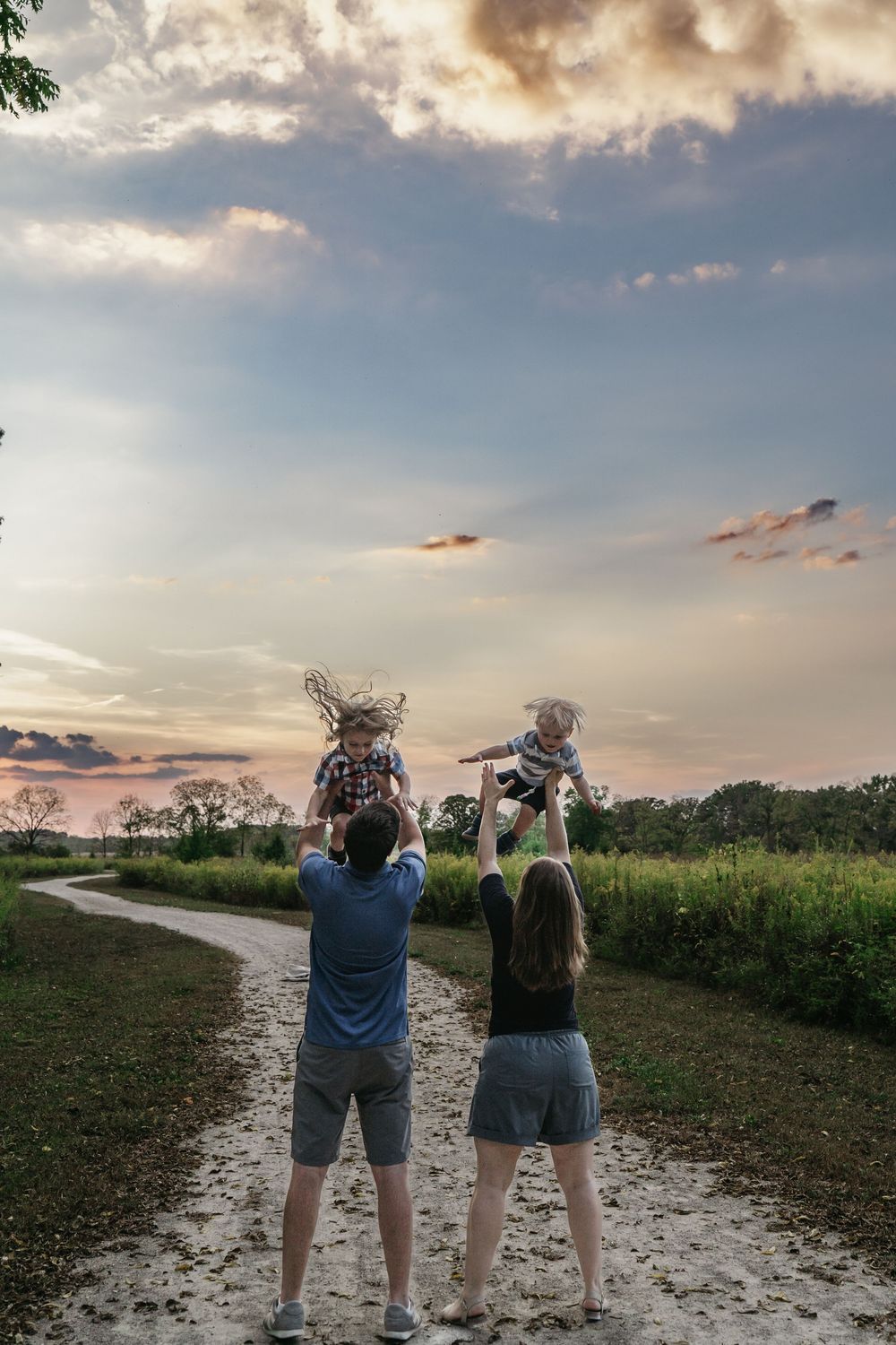 Two people holding children up in the air while standing on a path at sunset with colorful clouds in the sky for family photos in Ohio