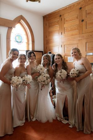 Bridesmaids in matching champagne dresses pose together holding white bouquets in a church hallway.