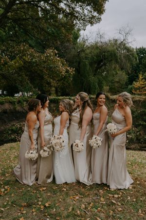 Wedding party of bridesmaids in elegant champagne gowns holding white bouquets outdoors.