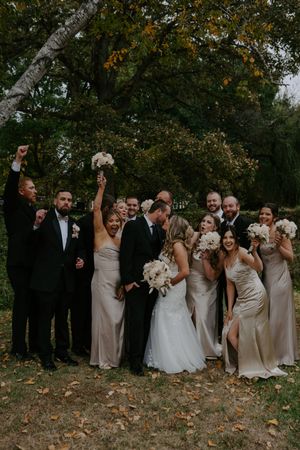 Wedding party celebrates energetically outdoors with raised arms and joyful expressions.