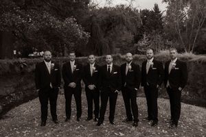 Groomsmen in black suits and ties stand in a row outdoors during an autumn wedding photoshoot.