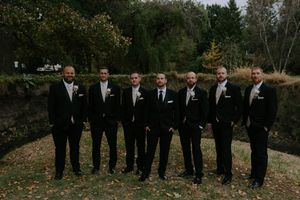 Groomsmen in black tuxedos pose together outdoors during fall wedding.