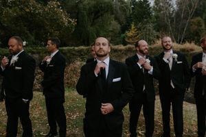 Row of groomsmen in black suits stand against autumn backdrop.