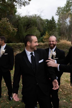 Wedding party members in black suits share laughs and candid moments in an outdoor fall setting.