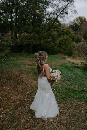 Bride in white lace gown poses outdoors on grassy hillside during fall season.