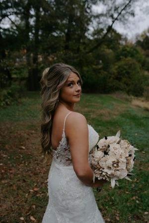 Bride holding neutral bouquet poses in lace dress against autumn landscape.
