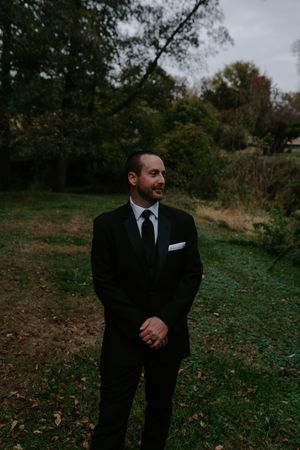 Groom in classic black tuxedo stands outdoors during fall wedding photoshoot.