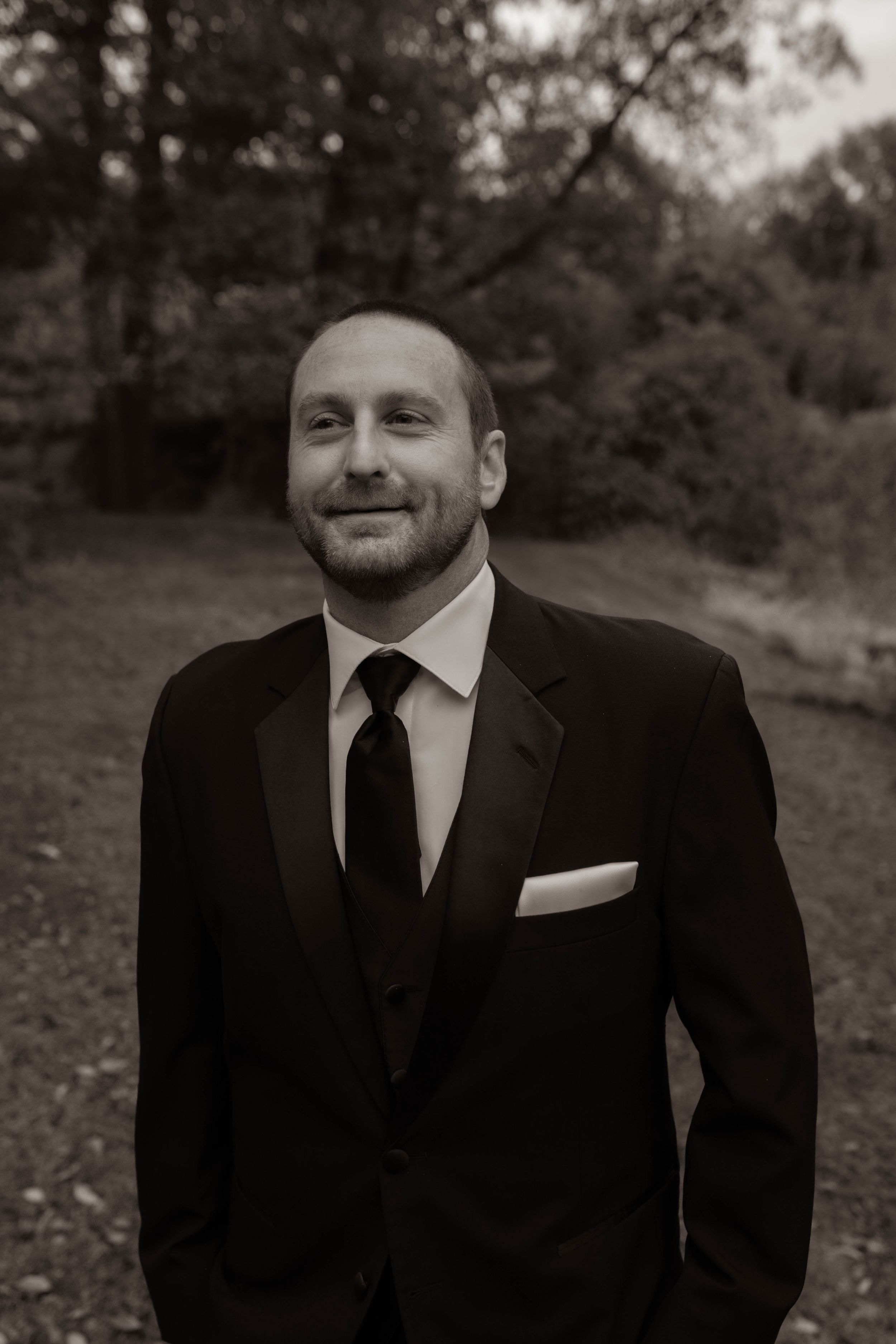 A groom in a classic black suit and tie poses outdoors in a black and white portrait.