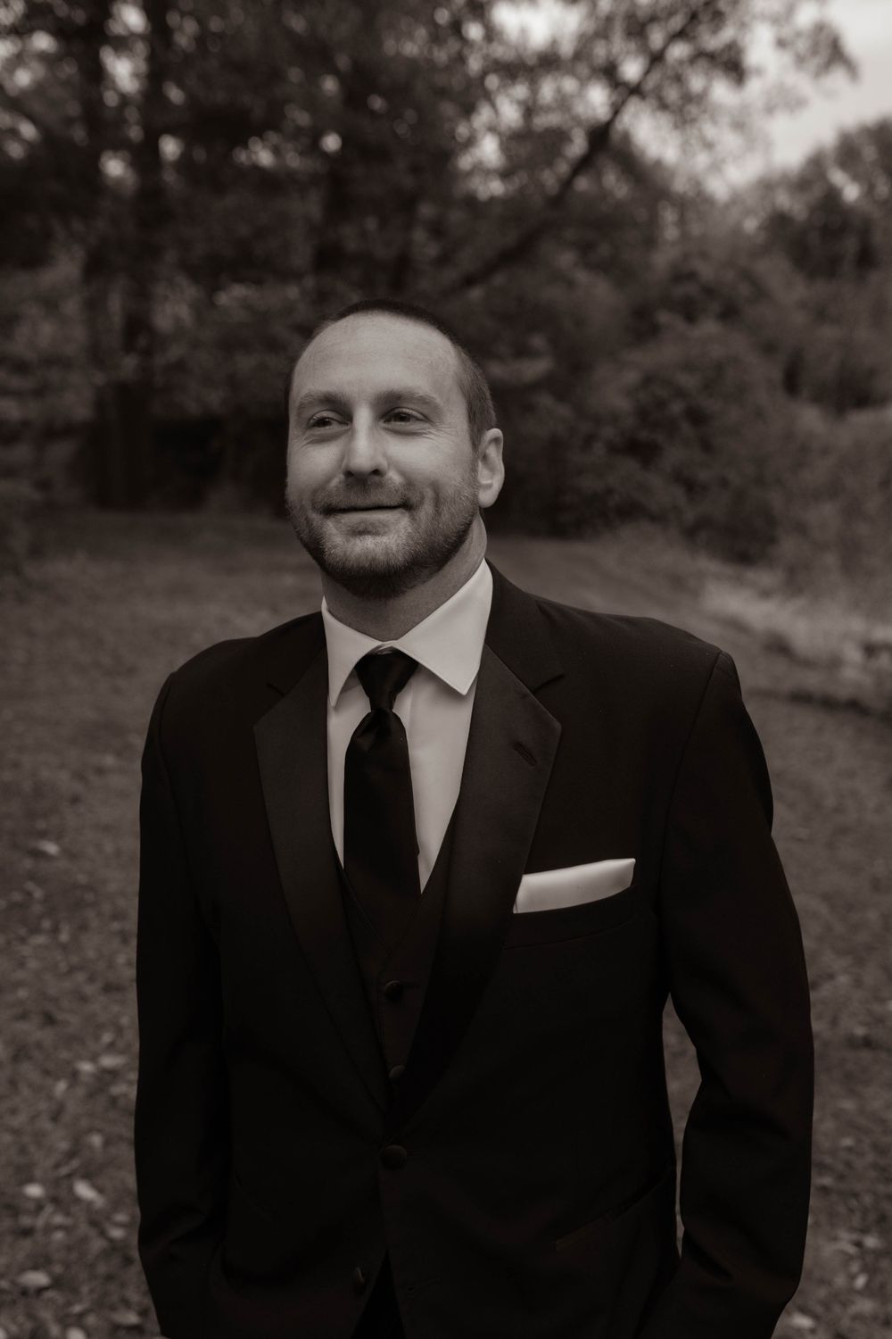 A groom in a classic black suit and tie poses outdoors in a black and white portrait.