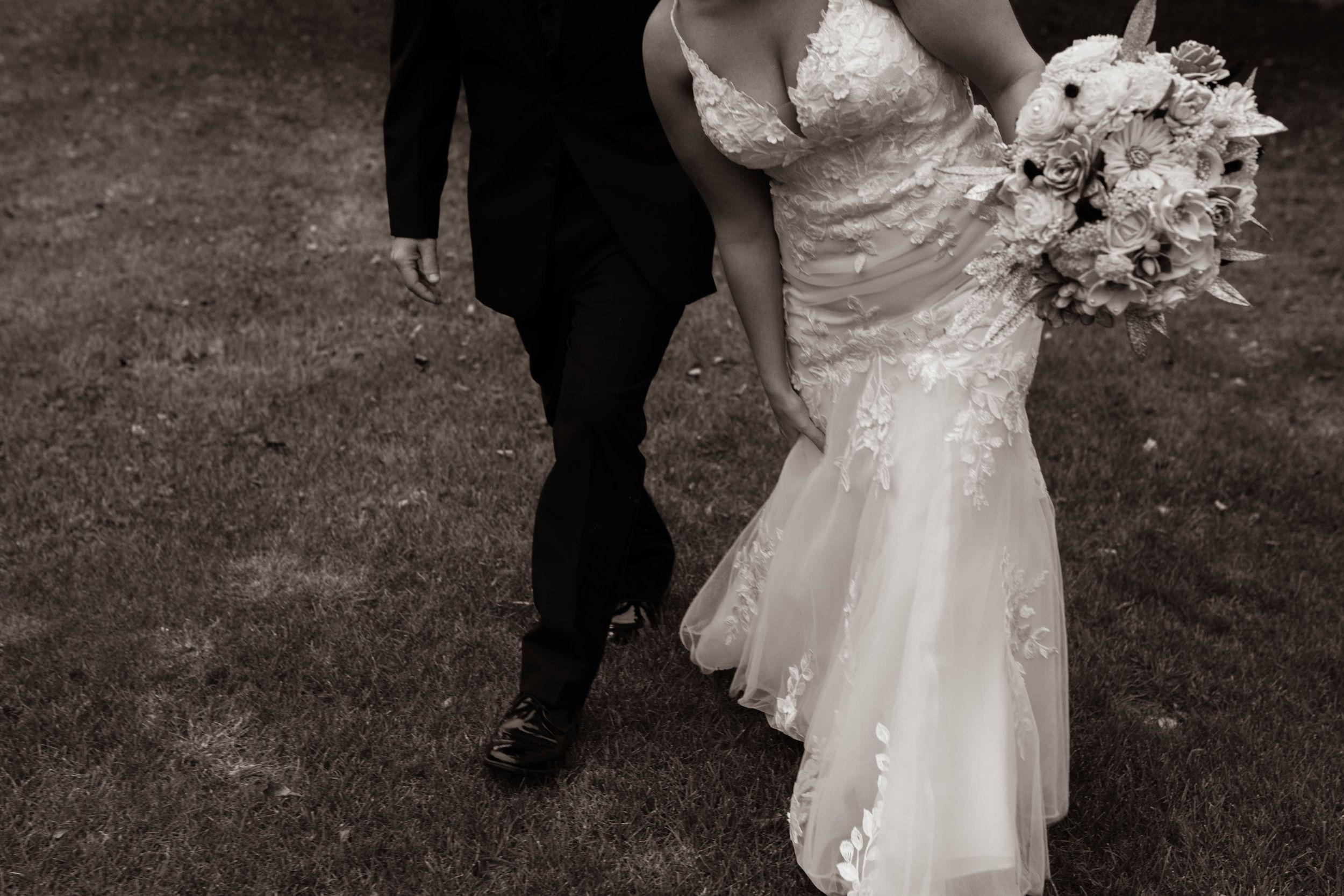 Black and white photo of newlyweds walking, showing bride's lace dress and groom's suit from waist down.