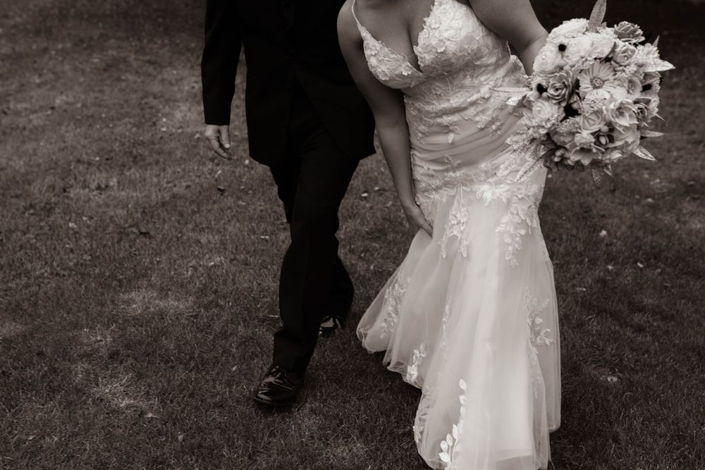 Black and white photo of newlyweds walking, showing bride's lace dress and groom's suit from waist down.