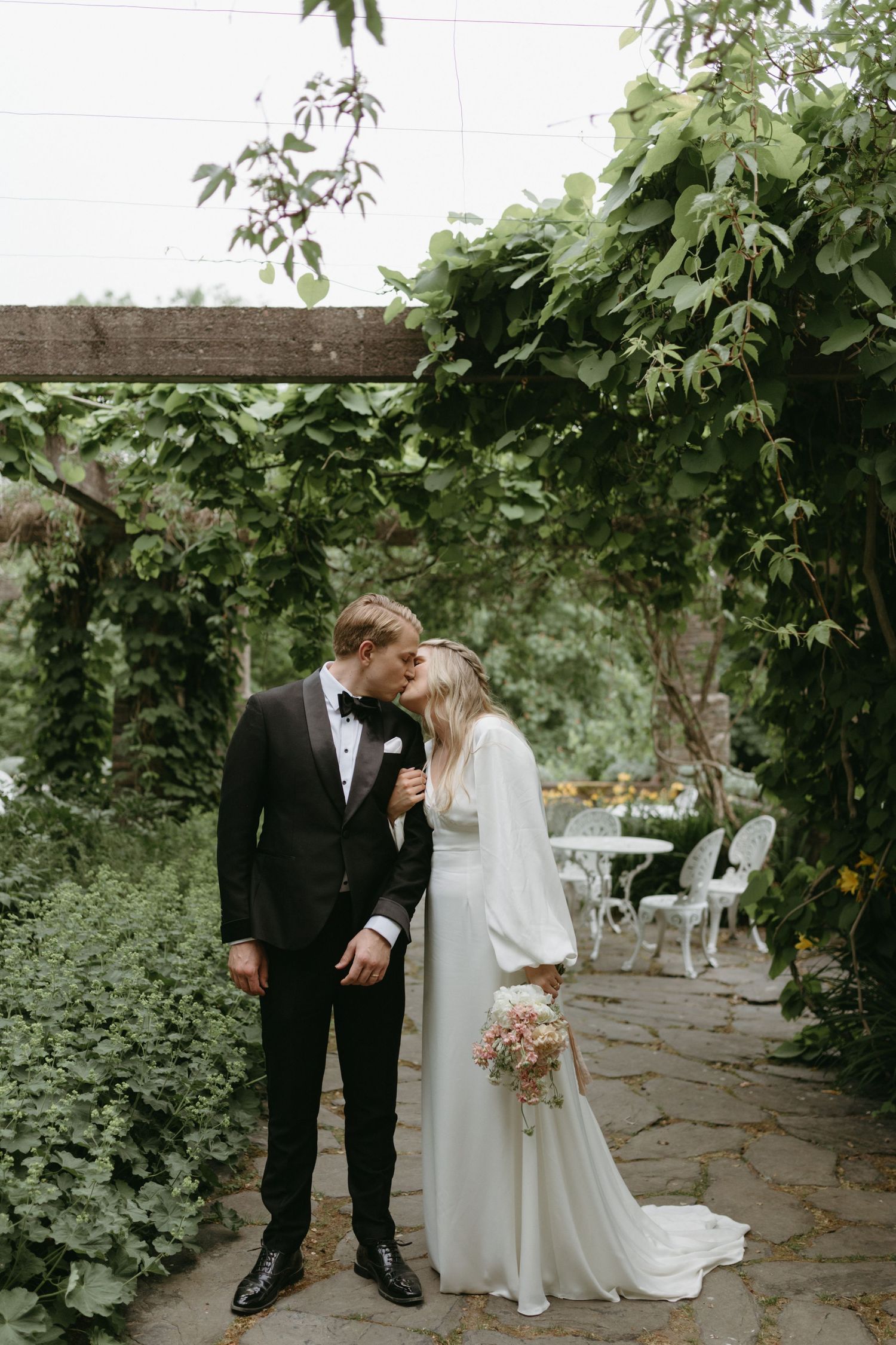 Elegant wedding guests gather outside a white mansion with blooming purple flowers in the garden.