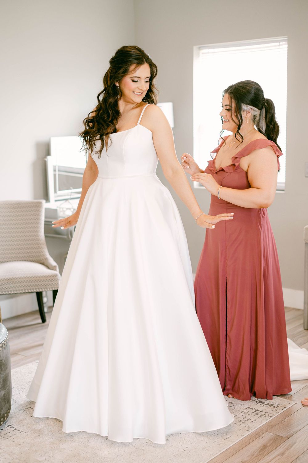 Two women in contrasting white and terracotta dresses stand together in a bright interior setting.