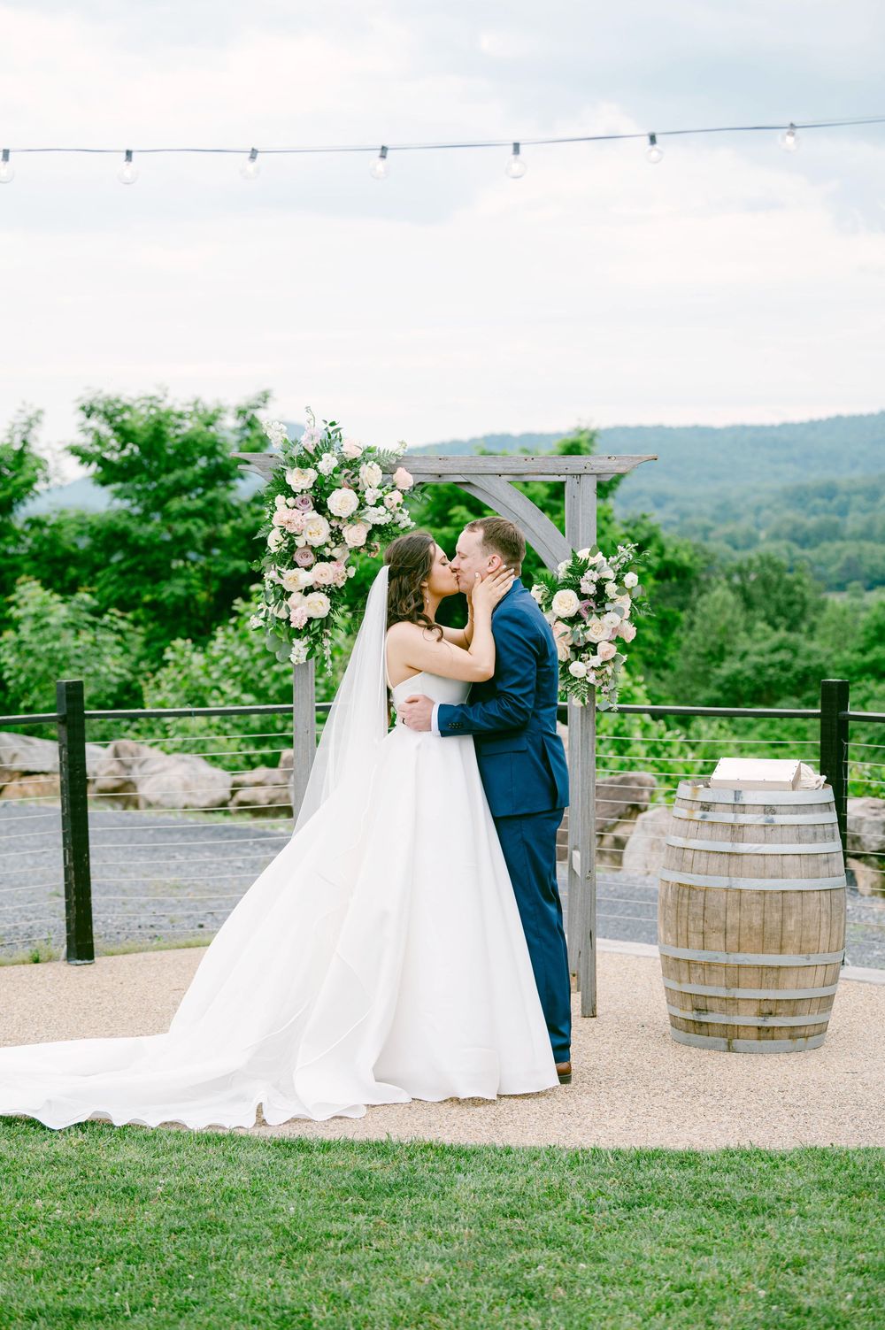 Elegant outdoor wedding couple shares romantic moments on a hilltop venue decorated with rustic wine barrels and greenery.