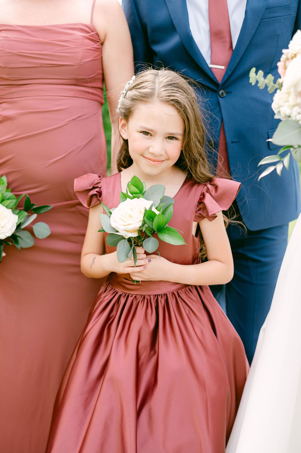 A young girl in a dusty rose dress holds a white flower while standing next to someone in a navy suit.