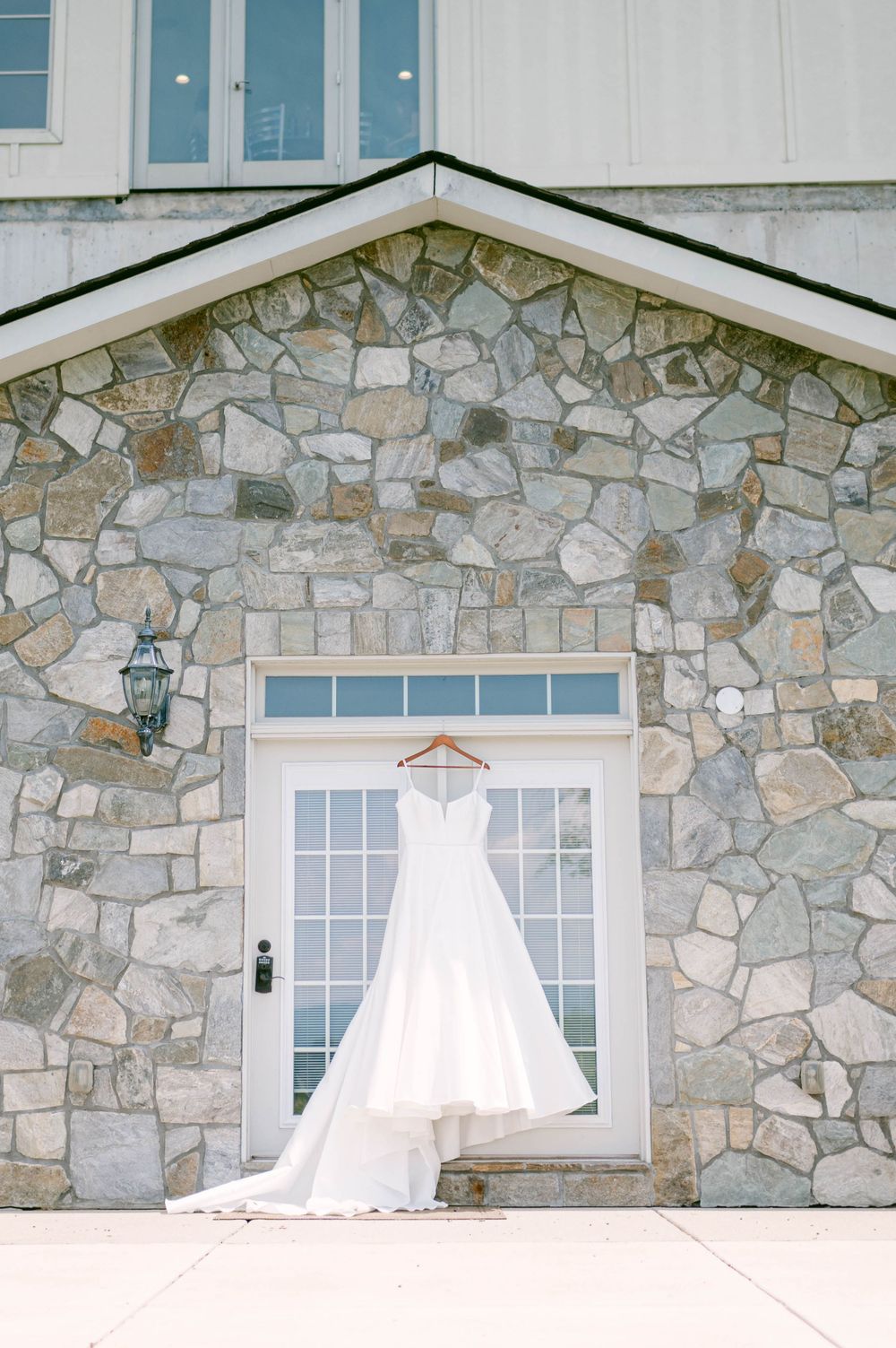 A wedding dress hangs in front of a rustic stone wall with windows, creating an elegant bridal preparation scene.