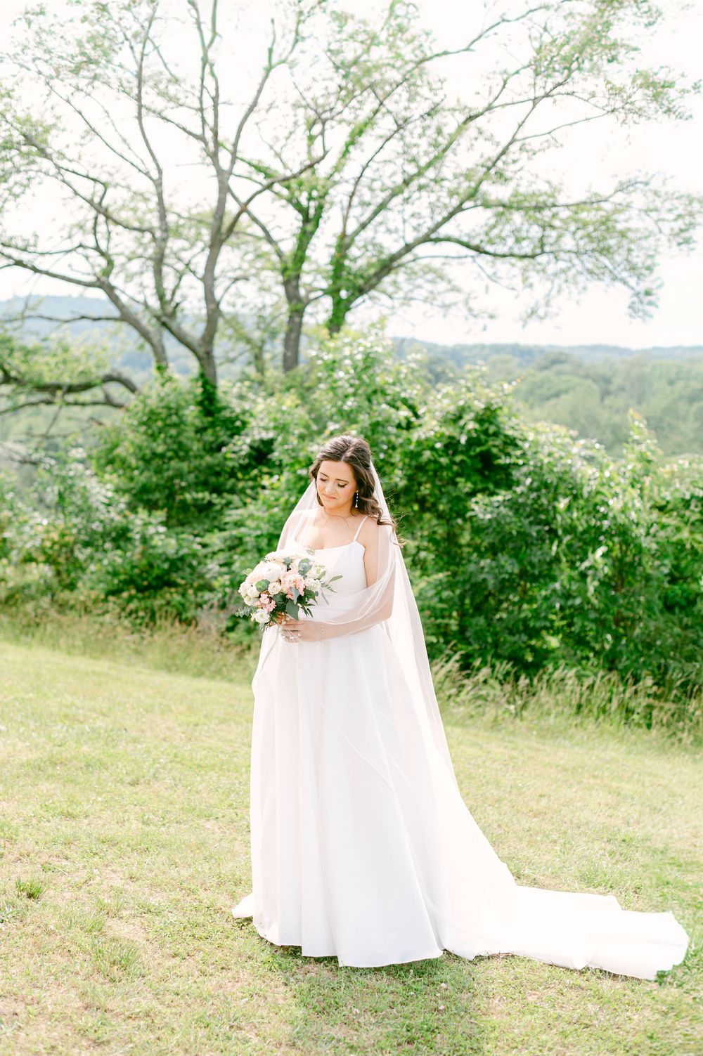 A bride in a flowing white gown with long veil holds a floral bouquet while standing on a hilltop with mountain views.