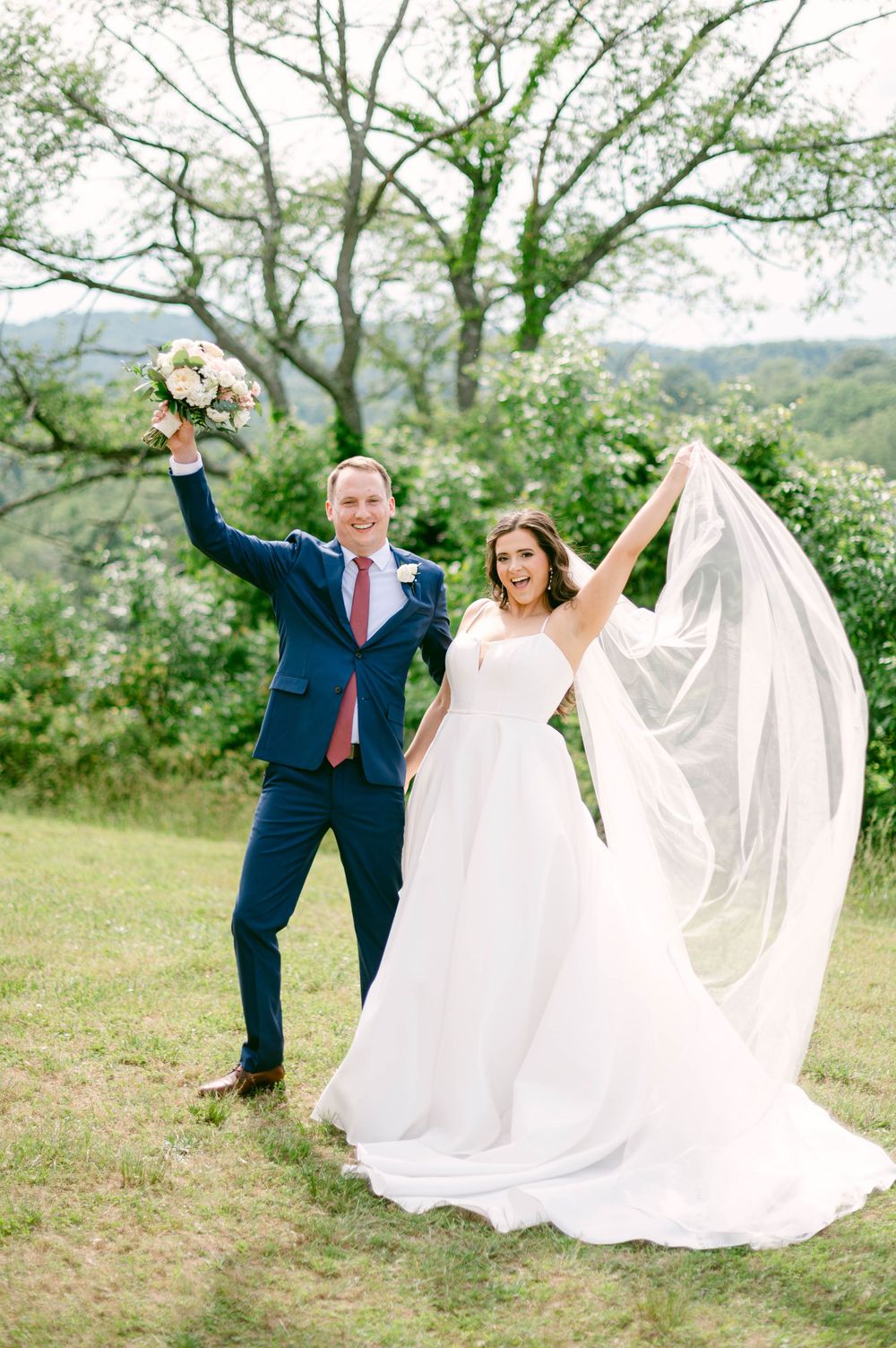 A couple in wedding attire playfully poses in a grassy field with trees, the bride's white dress flowing in the wind.
