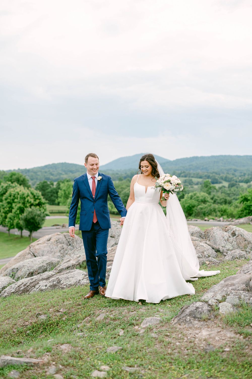 A bride and groom walk together on a scenic hilltop with rolling mountains in the background on their wedding day.