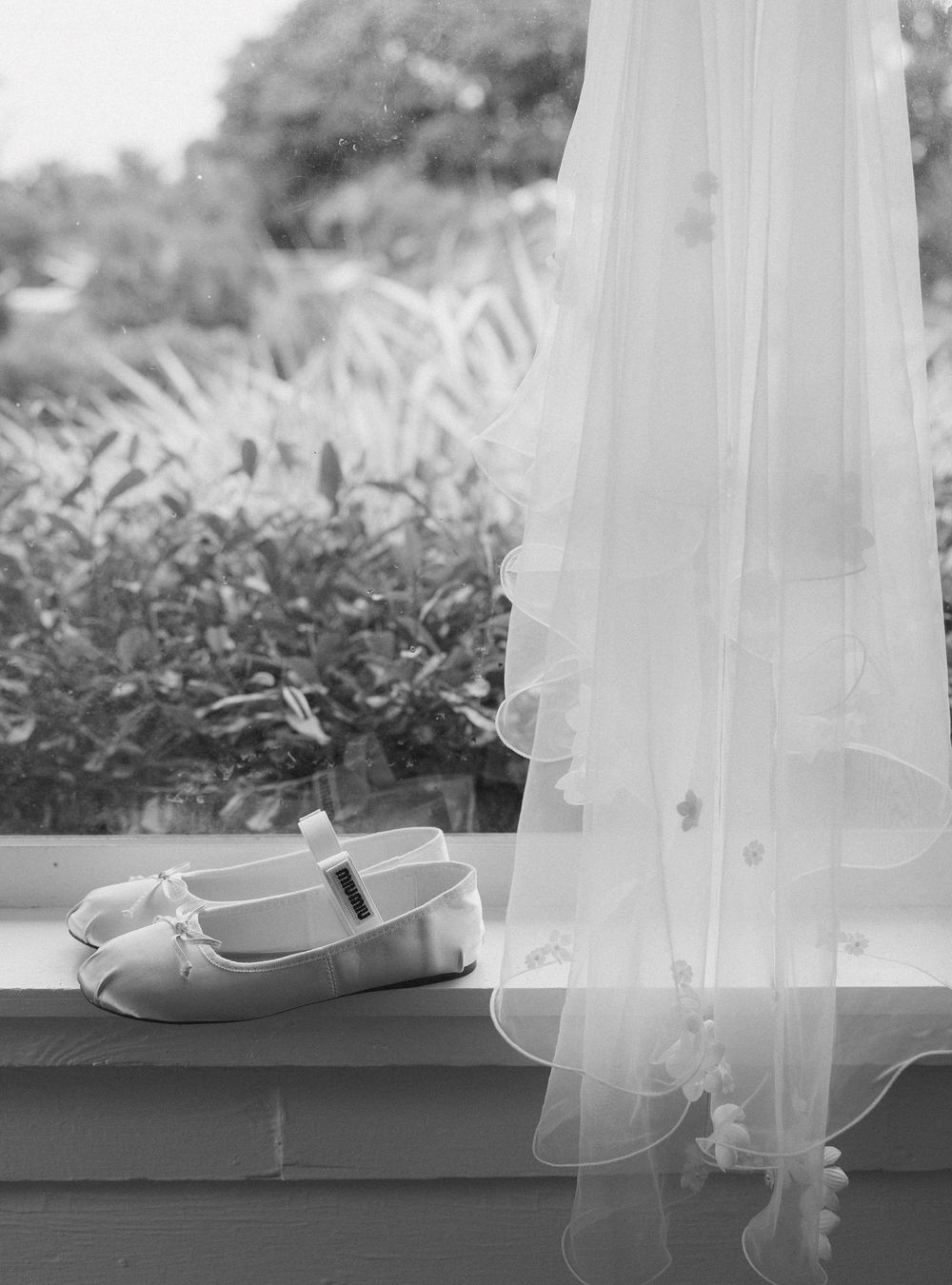 A pair of white ballet flats with delicate tulle ribbons sit on a dark surface in a black and white photograph.
