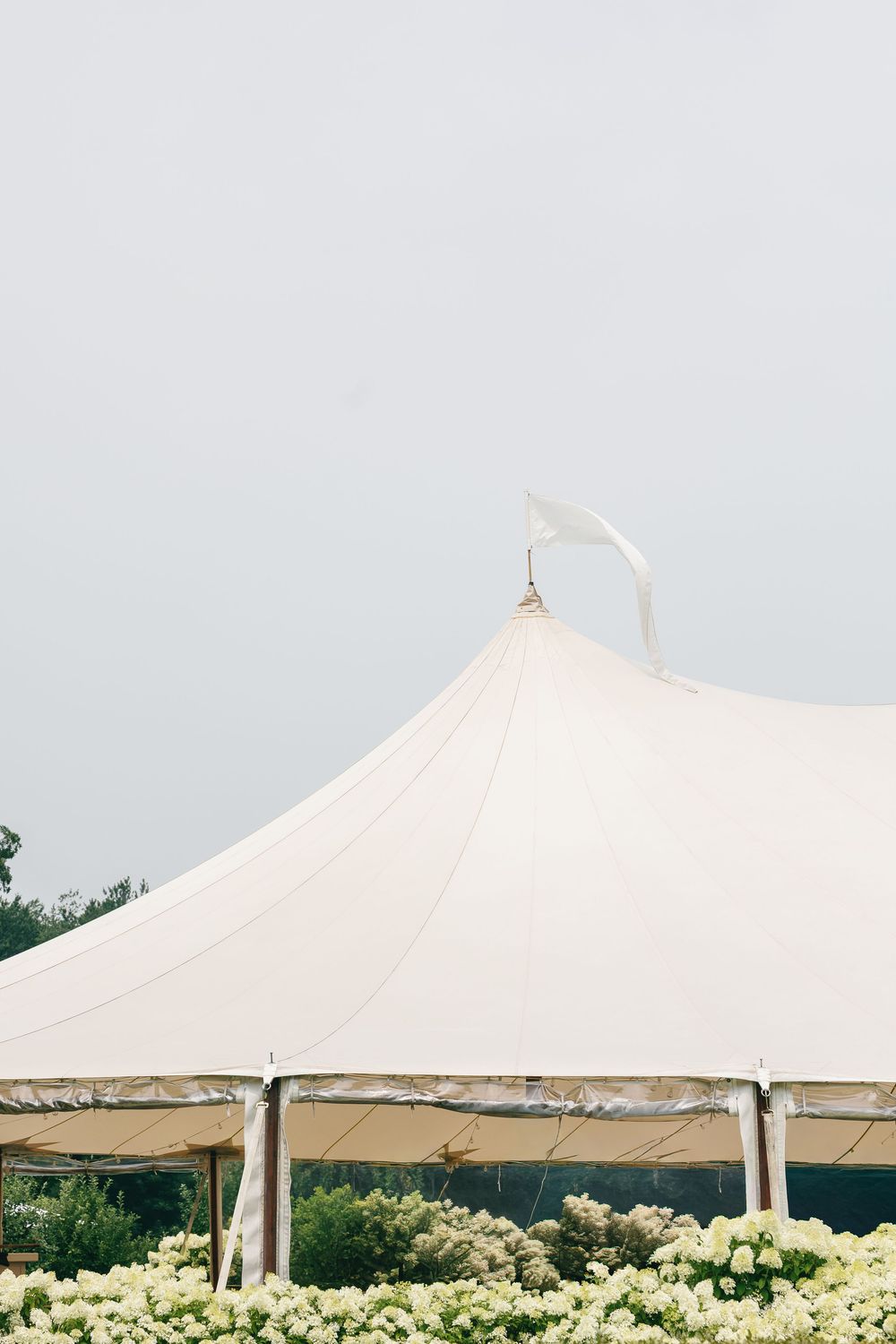 White sailcloth wedding tent surrounded by blooming white flowers against a soft gray sky.