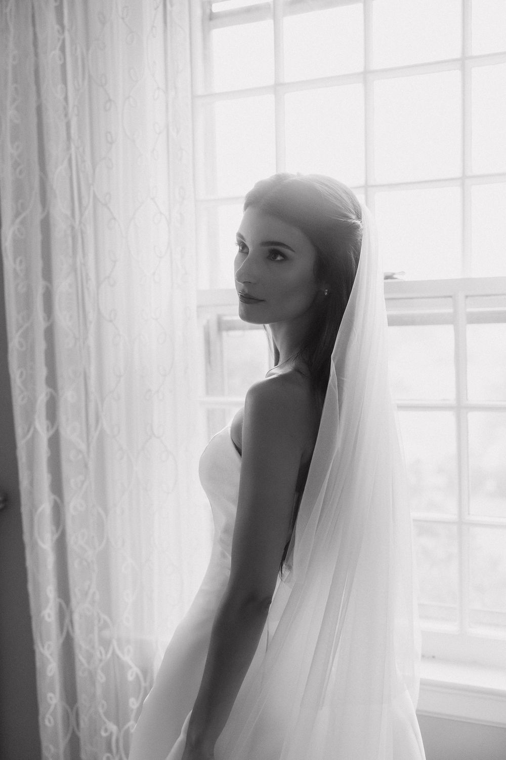Black and white portrait of a bride in a wedding veil standing by a bright window with lace curtains.