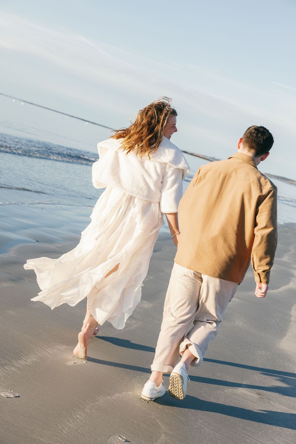 Couple in wedding attire walking hand in hand on a sandy beach with ocean waves in the background.