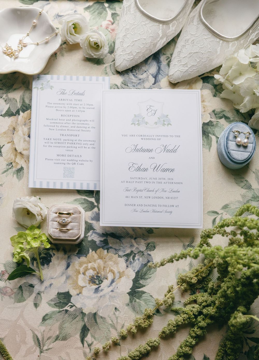 Wedding invitation suite with rings, floral details, lace shoes, and blue velvet ring box on floral backdrop.