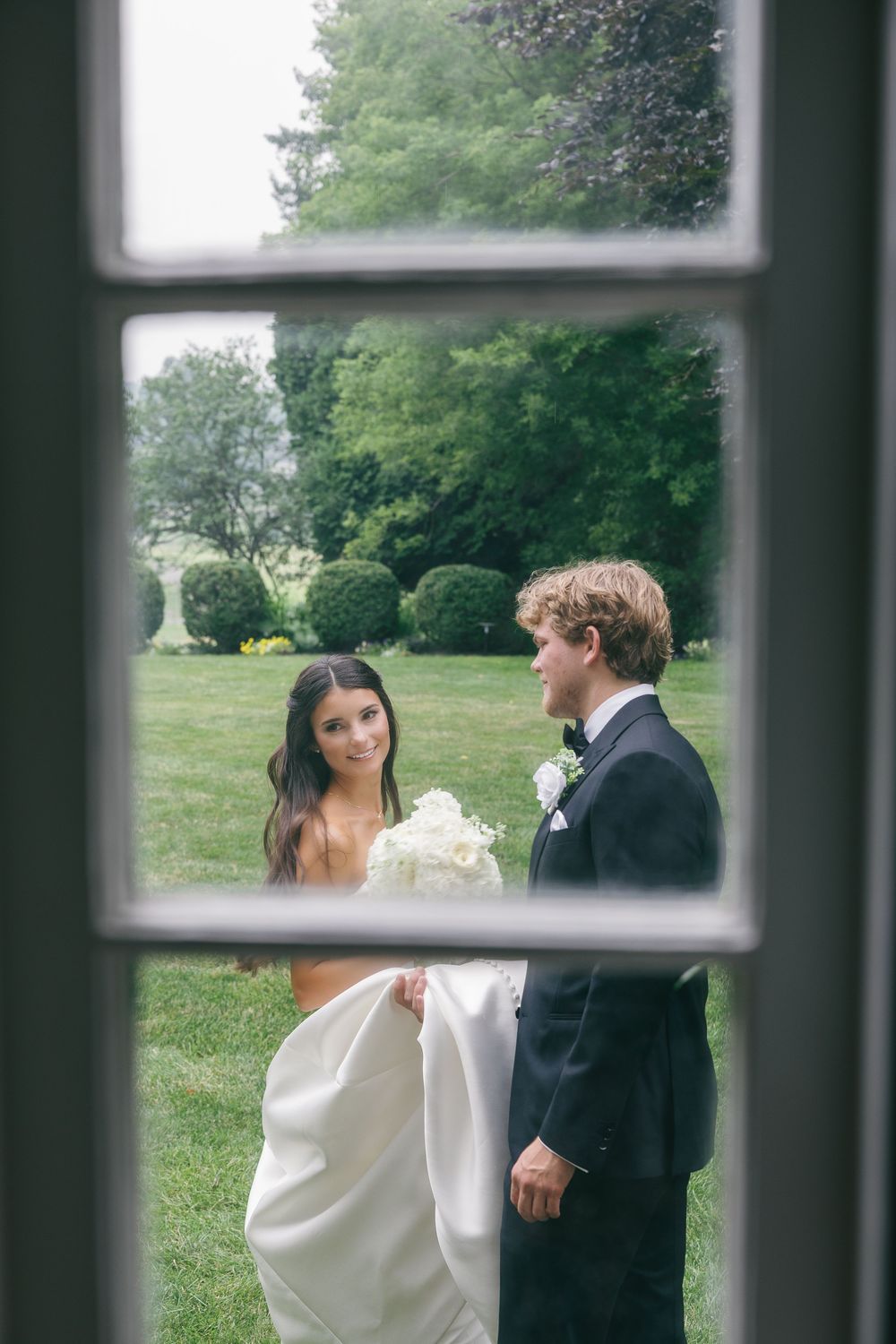 Bride and groom share a romantic moment in a lush garden, viewed through a window frame.