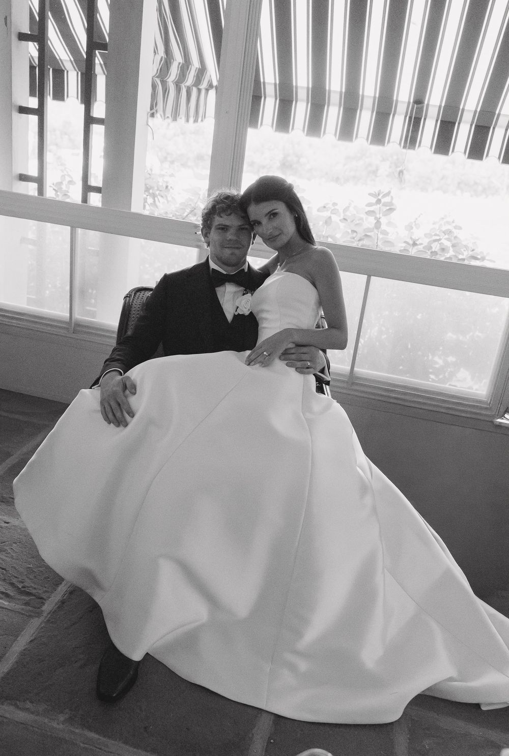 Black and white photo of a bride and groom posing together, bride in strapless gown, groom in tuxedo, seated indoors.
