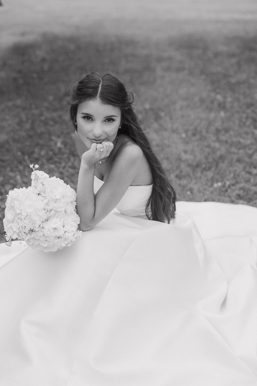 Black and white photo of a bride in a strapless gown sitting on grass, holding her bouquet of white flowers.